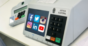 Imagem conceitual que exibe uma urna eletrônica de votação da Justiça Eleitoral do Brasil. Na tela digital da urna, estão sobrepostos vários ícones coloridos de redes sociais populares, como Facebook, X (antigo Twitter), Instagram, LinkedIn e YouTube. Ao lado da tela, observa-se o teclado numérico e os botões de confirmação, correção e voto em branco.