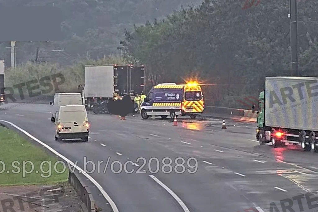 Cena de atendimento a um acidente de trânsito na Rodovia dos Bandeirantes captada por câmera de monitoramento, com ambulância da concessionária com girofles acesos, cones de sinalização na pista, caminhões e vans ao redor.