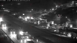 Captura de tela em preto e branco de uma câmera de monitoramento viário noturna da concessionária CCR AutoBan, identificada no topo como SP-348 (Rodovia dos Bandeirantes), quilômetro 70 no sentido Sul. A imagem mostra trecho de rodovia iluminado por postes de luz e faróis de veículos. Na pista da direita, observa-se uma fila de caminhões e carretas parados ou em tráfego lento. Na pista oposta, veículos trafegam com faróis acesos em direção ao observador. Há marcas d'água com o logotipo da ARTESP sobrepostas na imagem.