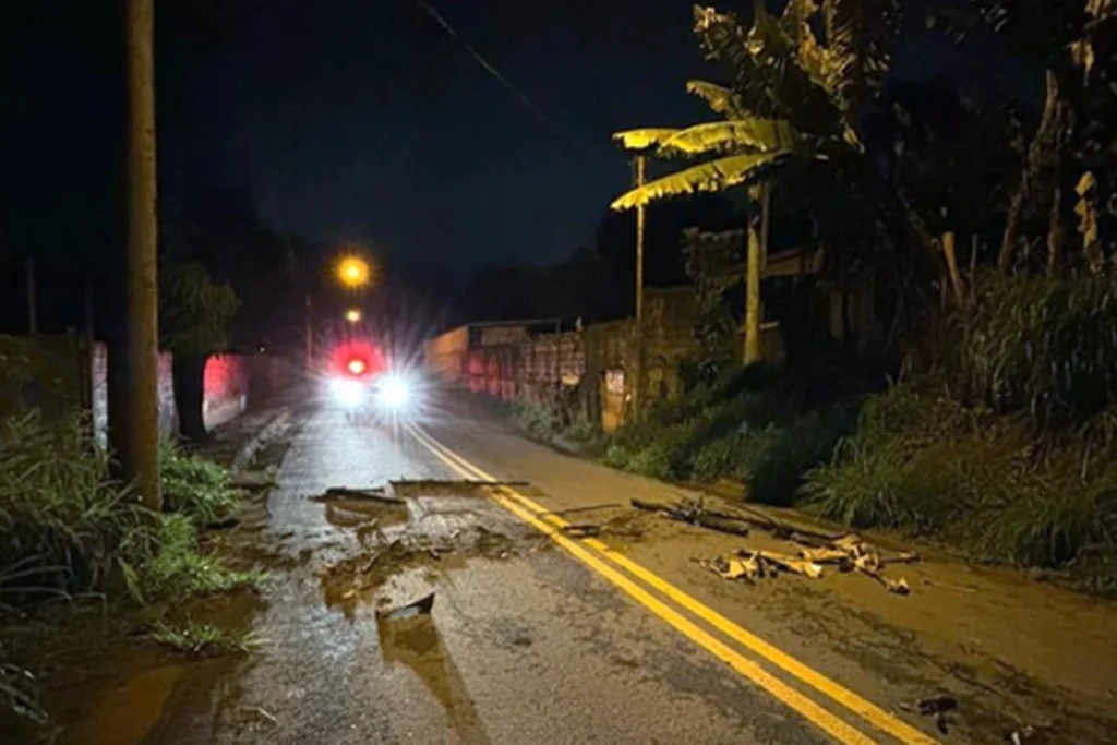 Fotografia noturna de uma via de mão dupla asfaltada com faixa central amarela dupla, onde há acúmulo de lama, sujeira e pedaços de galhos espalhados sobre o asfalto. Ao fundo, no centro da pista, vê-se uma viatura com os faróis e o giroflex vermelho acesos. Nas laterais da via há postes de iluminação, muros e vegetação densa, incluindo uma bananeira à direita.