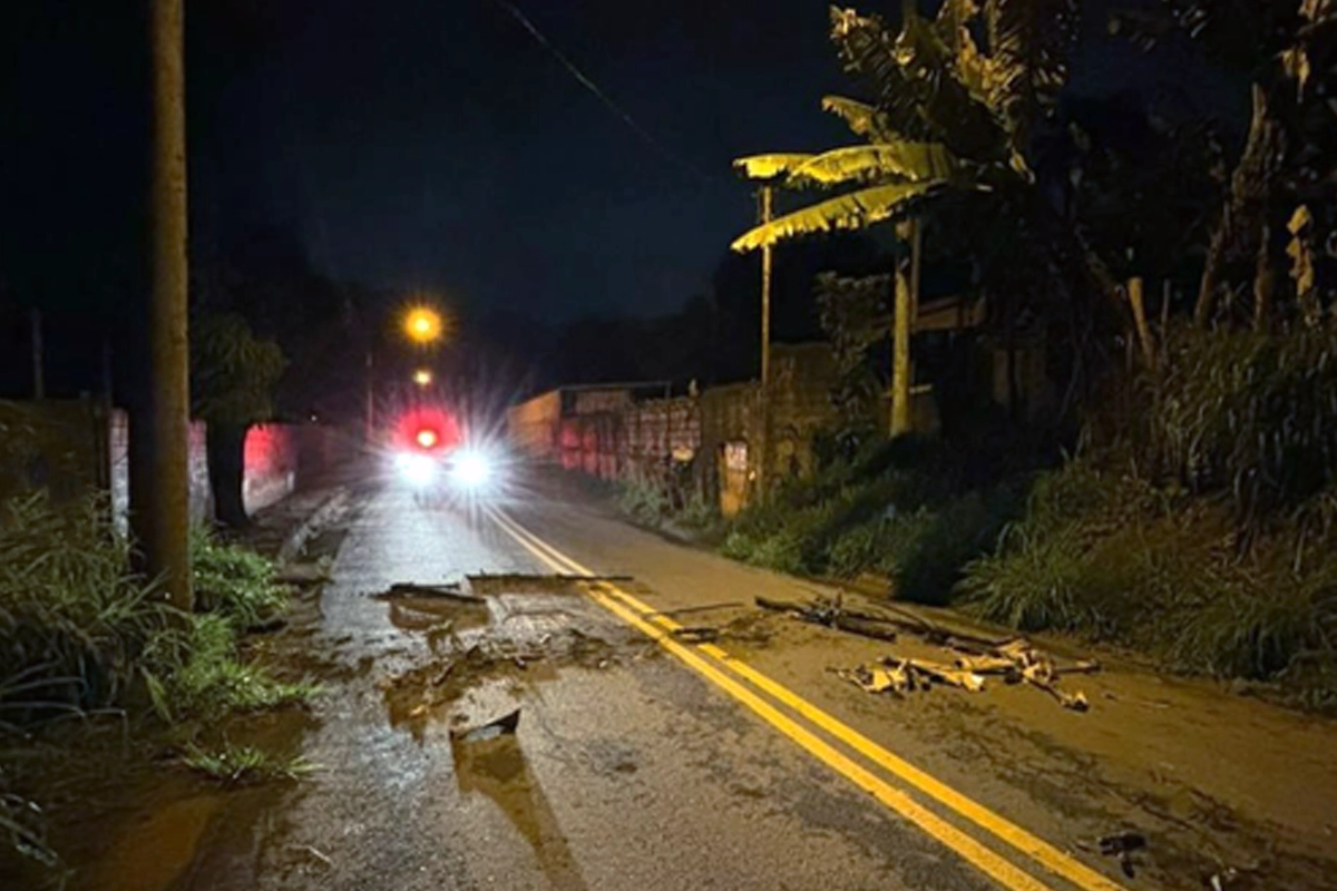 Fotografia noturna de uma via de mão dupla asfaltada com faixa central amarela dupla, onde há acúmulo de lama, sujeira e pedaços de galhos espalhados sobre o asfalto. Ao fundo, no centro da pista, vê-se uma viatura com os faróis e o giroflex vermelho acesos. Nas laterais da via há postes de iluminação, muros e vegetação densa, incluindo uma bananeira à direita.