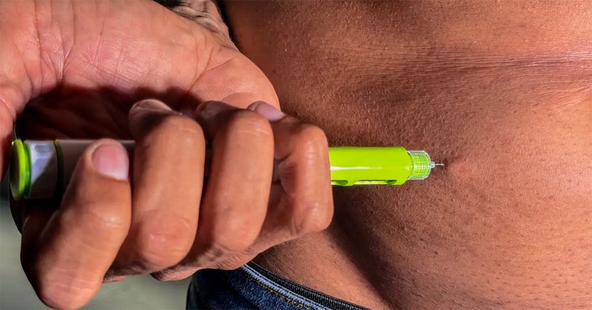 Fotografia em close-up que mostra as mãos de uma pessoa segurando uma caneta injetora verde e aplicando o medicamento na pele da região abdominal. A agulha fina do dispositivo está encostada na pele no momento da aplicação subcutânea.