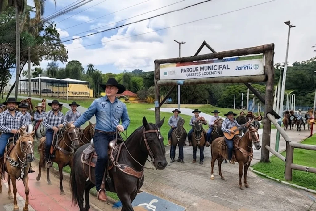 Grupo de cavaleiros trajando chapéus e camisas de estilo sertanejo montados em cavalos na entrada do Parque Municipal de Jarinu. Um dos cavaleiros, em primeiro plano sobre um cavalo escuro, acena enquanto lidera o grupo diante de um pórtico de madeira.