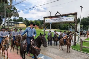 Grupo de cavaleiros trajando chapéus e camisas de estilo sertanejo montados em cavalos na entrada do Parque Municipal de Jarinu. Um dos cavaleiros, em primeiro plano sobre um cavalo escuro, acena enquanto lidera o grupo diante de um pórtico de madeira.