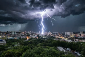 Raio brilhante caindo sobre uma cidade sob céu escuro de tempestade, com luzes urbanas acesas ao fundo e uma vasta área verde de árvores em primeiro plano.