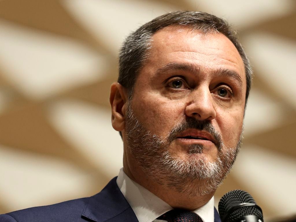 Primeiro-plano do diretor-geral da Polícia Federal brasileira, Andrei Rodrigues, vestindo terno azul e barba grisalha/castanha, falando ao microfone, com fundo geométrico em tons beges.