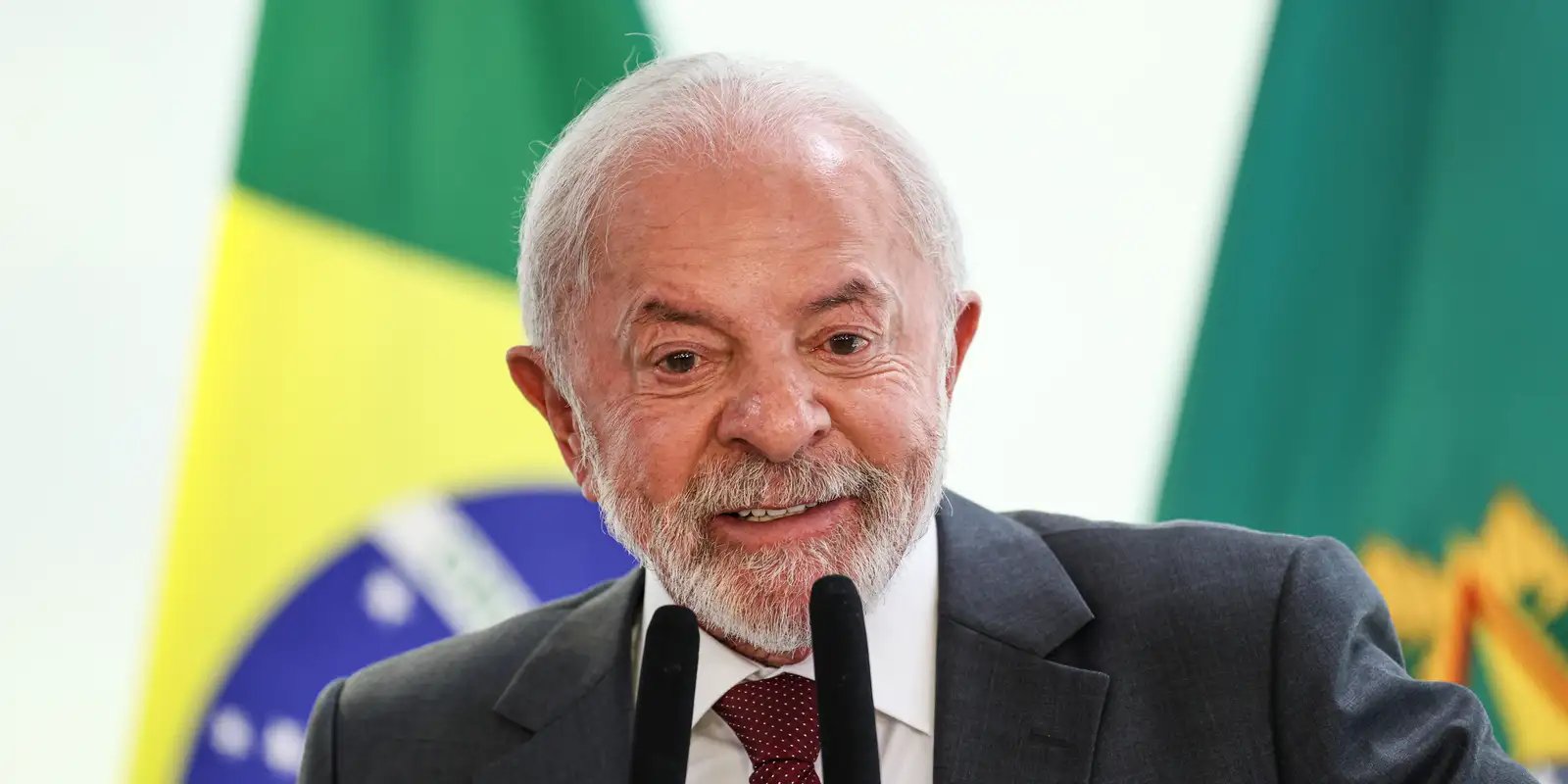 O presidente Luiz Inácio Lula da Silva sorrindo enquanto discursa ao microfone, vestindo terno cinza, camisa branca e gravata bordô, com a bandeira do Brasil ao fundo.