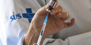 Primeiro-plano das mãos de um profissional de saúde vestindo jaleco com o logotipo do SUS, aspirando a dose de uma vacina de um pequeno frasco de vidro usando uma seringa com agulha.