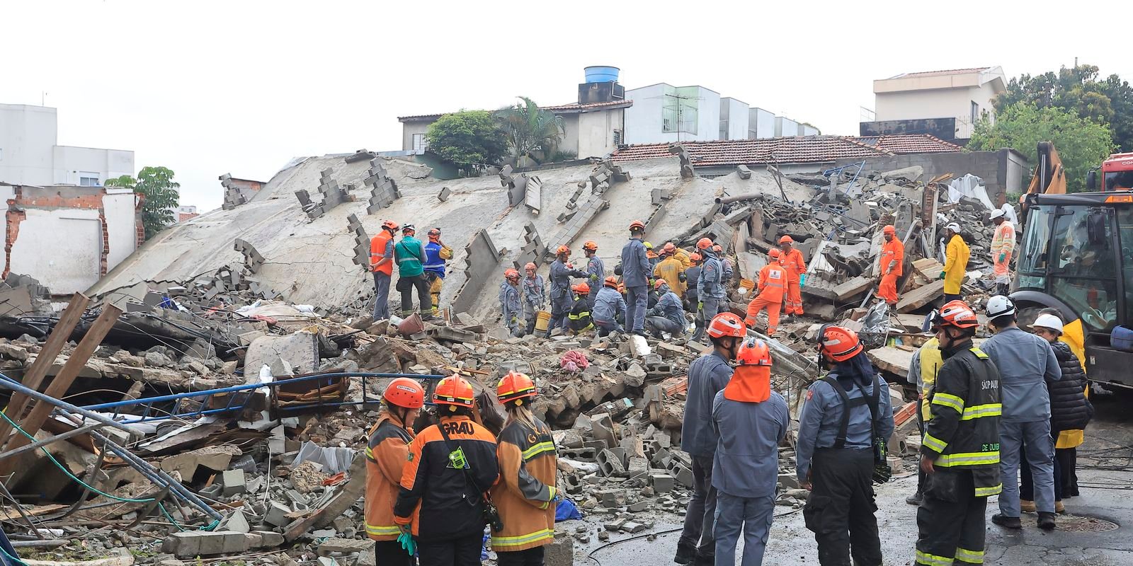 Fotografia de uma cena de resgate em um local de desabamento urbano. No centro e ao fundo, vê-se uma grande estrutura de concreto e tijolos completamente destruída. Diversos socorristas, bombeiros e profissionais do GRAU (Grupo de Resgate e Atenção às Urgências) usando capacetes de segurança e uniformes de proteção de cores laranja, azul e amarelo trabalham sobre os escombros e conversam no primeiro plano. À direita, parte de uma máquina escavadeira está visível.