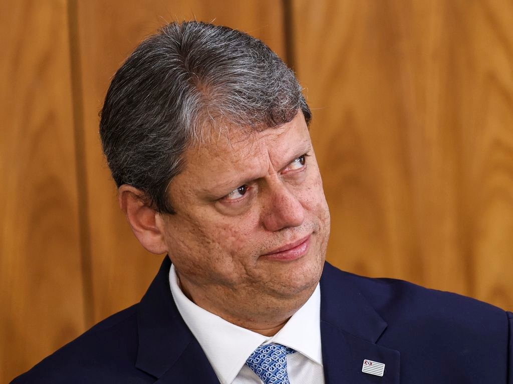 Tarcísio de Freitas vestindo terno azul-escuro, camisa branca e gravata azul estampada, com um broche da bandeira do Estado de São Paulo na lapela e um painel de madeira ao fundo.