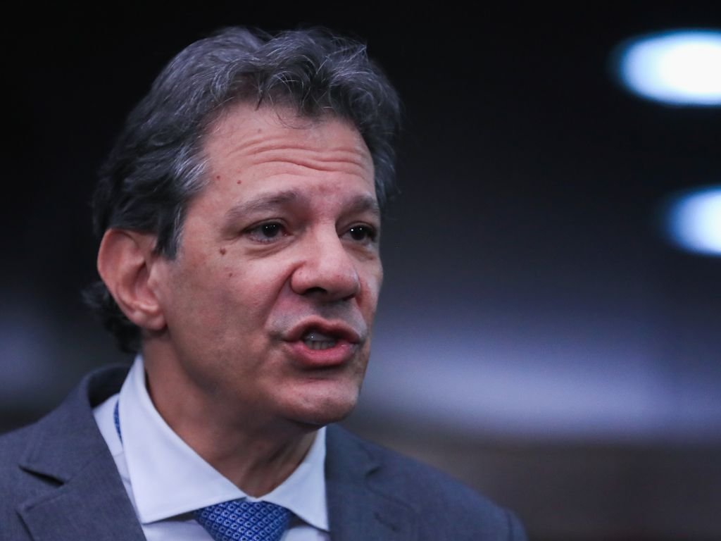 Fernando Haddad vestindo terno cinza, camisa branca e gravata azul estampada enquanto fala em um evento com fundo escuro.