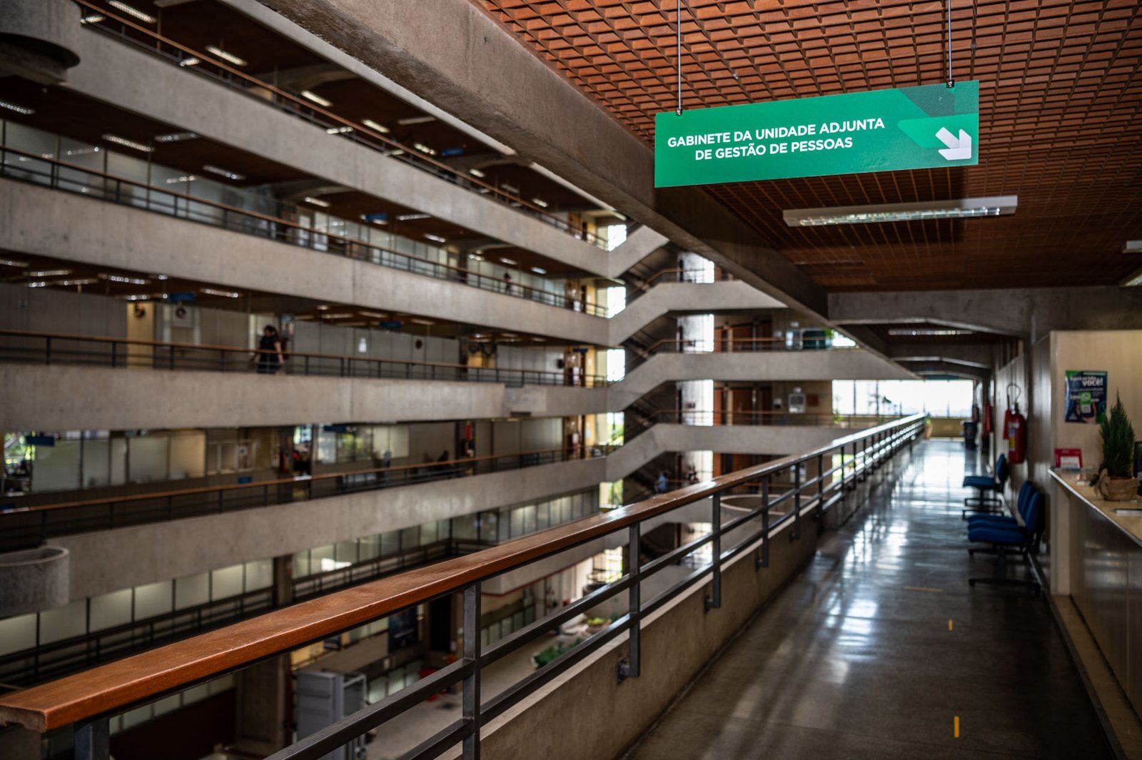 Placa de sinalização verde suspensa no teto, em um corredor interno do prédio da Prefeitura de Jundiaí com arquitetura em concreto aparente e múltiplos andares.