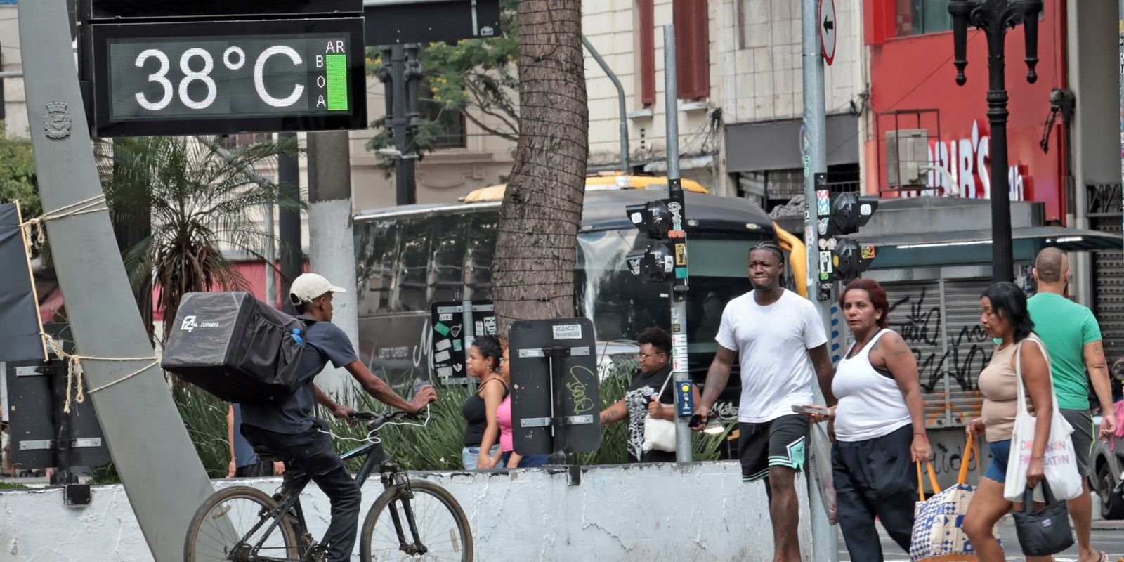 Fotografia de uma rua movimentada em um dia de calor intenso. No canto superior esquerdo, um termômetro digital de rua exibe a temperatura de 38°C. Em primeiro plano, pedestres caminham pela calçada vestindo roupas leves e um entregador de aplicativo anda de bicicleta transportando uma mochila térmica nas costas. Ao fundo, veem-se ônibus e outros veículos no trânsito urbano
