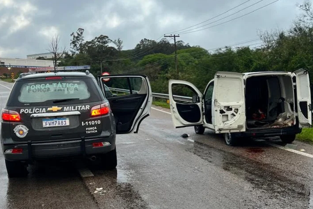 Fotografia de uma ocorrência policial em na rodovia D. Gabriel, sob céu nublado. No primeiro plano, à esquerda, vê-se a traseira de uma viatura escura da Força Tática da Polícia Militar de São Paulo com a porta do passageiro aberta. À direita, uma furgoneta/van de carga branca está parada com as portas dianteiras e traseiras abertas, revelando o compartimento de carga vazio. O asfalto está molhado e o local é ladeado por vegetação.
