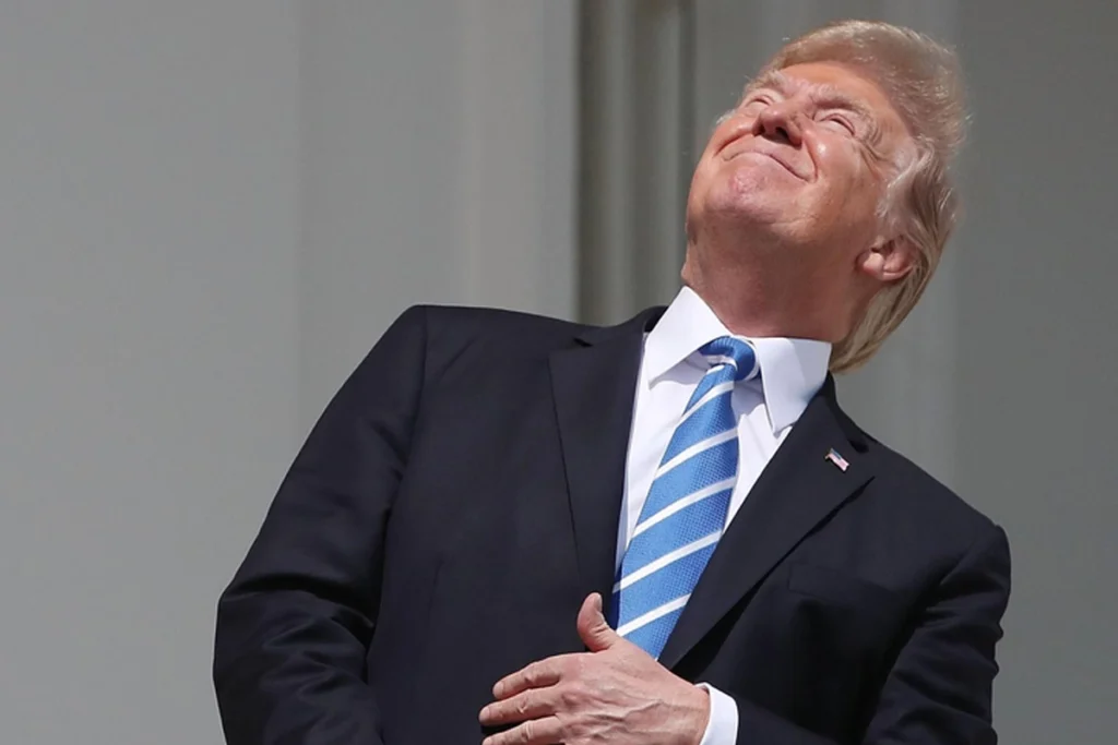 Donald Trump vestindo terno escuro, camisa branca e gravata listrada em azul e branco, olhando para o céu com a cabeça inclinada para trás e a mão direita sobre o peito enquanto está na sacada da Casa Branca