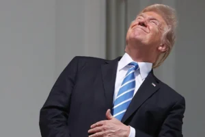 Donald Trump vestindo terno escuro, camisa branca e gravata listrada em azul e branco, olhando para o céu com a cabeça inclinada para trás e a mão direita sobre o peito enquanto está na sacada da Casa Branca