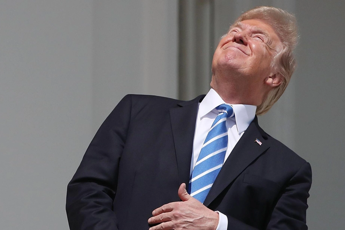Donald Trump vestindo terno escuro, camisa branca e gravata listrada em azul e branco, olhando para o céu com a cabeça inclinada para trás e a mão direita sobre o peito enquanto está na sacada da Casa Branca
