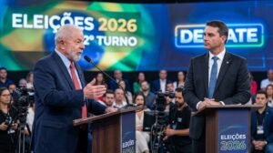 Uma fotografia em estilo editorial com qualidade de alta resolução mostra Luiz Inácio Lula da Silva e Flávio Bolsonaro em um debate de TV para o segundo turno das eleições presidenciais de 2026. O plano médio horizontal captura ambos os homens em pé atrás de pódios de madeira escura e voltados para a esquerda em relação ao plano de visão, ocupando o centro da moldura. À esquerda, Lula está com um terno azul-marinho, camisa azul-clara e gravata vermelha padronizada, e fala ao microfone do pódio com as mãos gestualizadas. À direita, Flávio está com um terno azul-marinho, camisa branca e gravata azul padronizada, e ouve com os braços cruzados no pódio. No fundo, há um grande painel de LED com gráficos abstratos em azul e verde com o texto em branco: "ELEIÇÕES 2026" e "2º TURNO". Um painel menor à direita, mais desfocado, tem o texto: "DEBATE". Uma plateia sentada e operadores de câmera são vistos desfocados atrás dos homens. A iluminação é de estúdio.