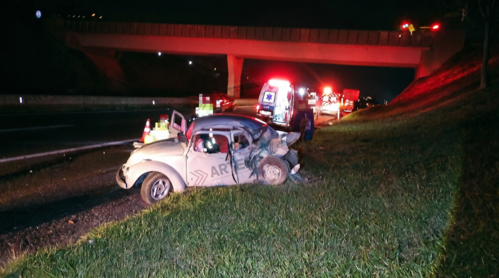 Fotografia noturna de um acidente de trânsito em rodovia sob um viaduto. Em primeiro plano, no gramado ao lado do acostamento, vê-se um Volkswagen Fusca claro com a parte traseira e a capota completamente amassadas e destruídas. Ao fundo, uma ambulância com giroflex ligado e veículos de resgate realizam o atendimento no local. A imagem traz a marca d'água da ARTESP